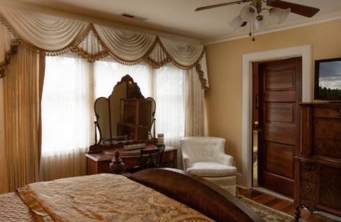 Luxurious bedroom in shades of gold and ivory with dresser and mirror, sitting chair and large chest with TV